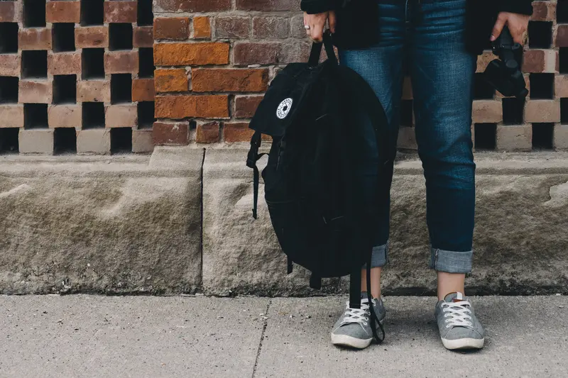 Pieds de jeune avec sac à dos à la main devant un mur d'établissement scolaire dans la rue, cour d'école